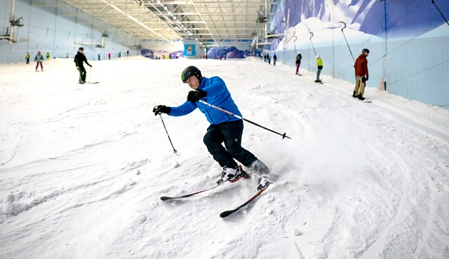 Man in a blue jacket skiing in the snow, there are others skiing in the background.