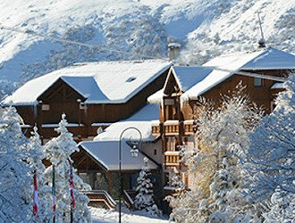 A mountain ski chalet with snow on the roof, set in snowy landscape amongst trees.