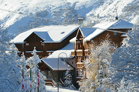 A mountain ski chalet with snow on the roof, set in snowy landscape amongst trees.