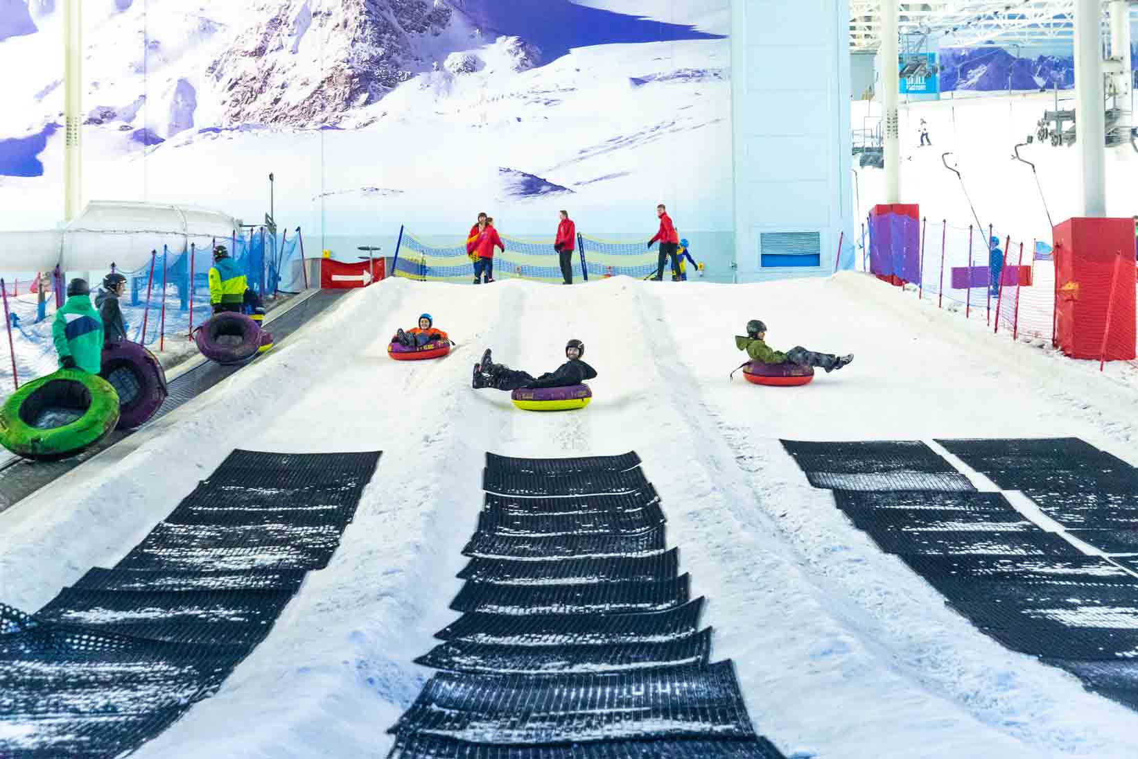 Three people racing down the chill factore snowy slope in inflatable donuts, wearing helmets