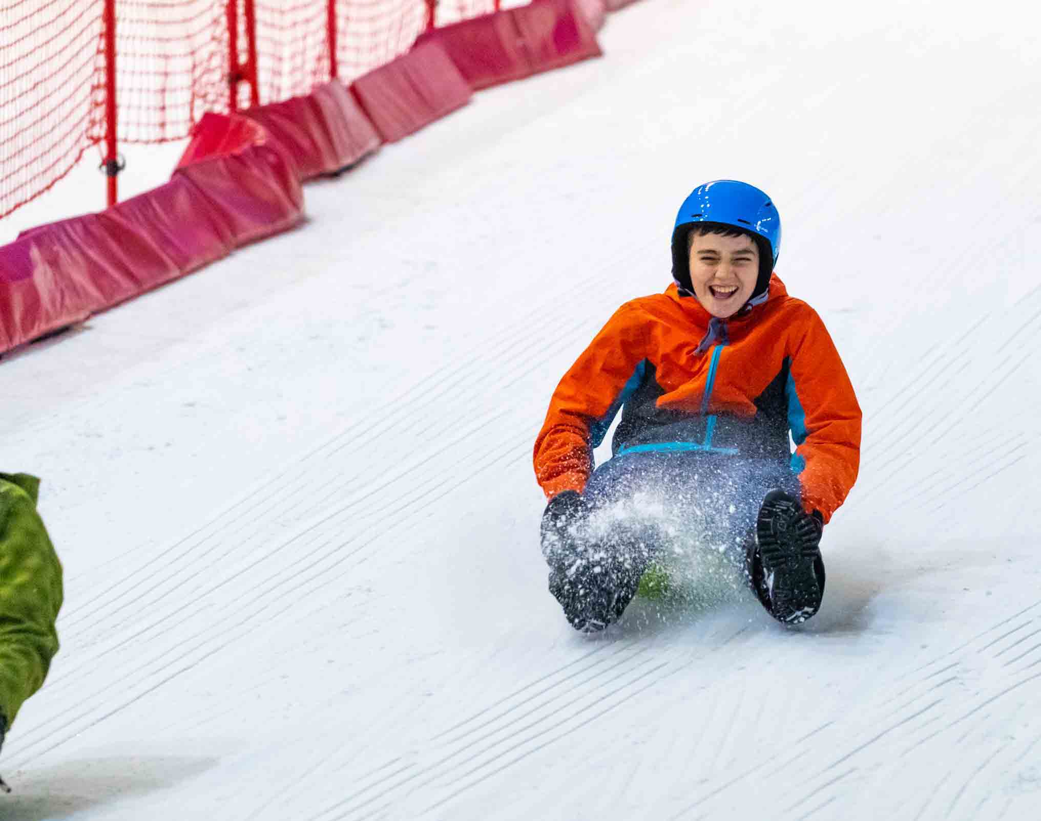 A child wearing a red jacket and blue helmet, sliding on a sledge down a snowy slope